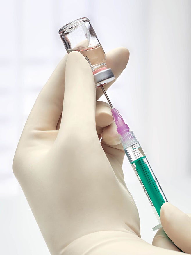 Close-up of a gloved hand drawing solution from a transparent vial with a syringe, illustrating sterile drug preparation with syringe and vial.