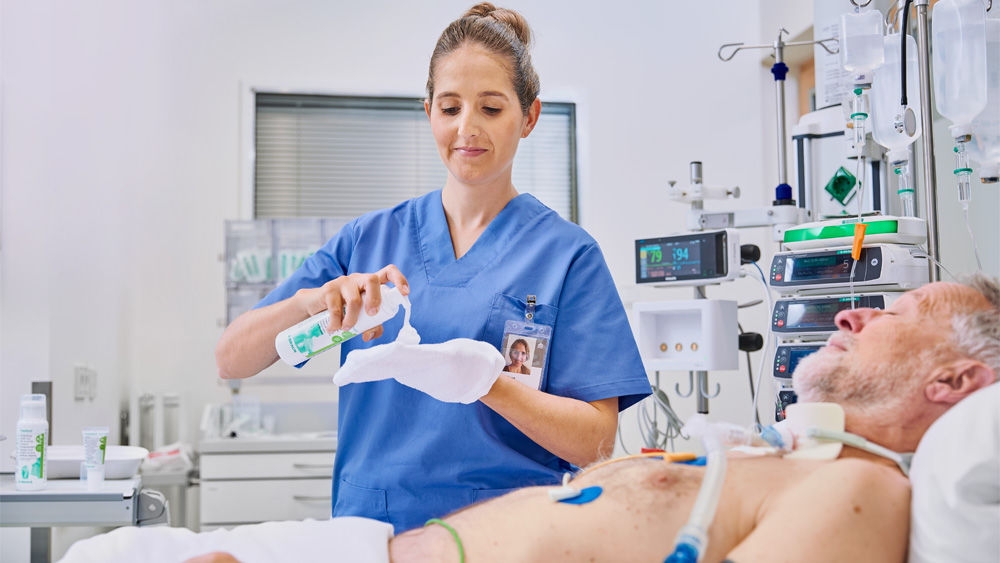 female nurse holding Prontoderm foam in hospital in front of an elderly patient lying in bed and applying it with a glove