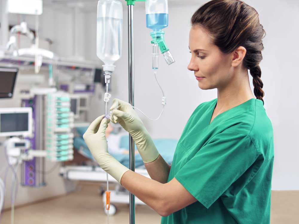 A nurse in a green coat hangs up an IV bottle in an ICU room. Two Ecoflac plus bottles from B. Braun hang on the stand and are connected.