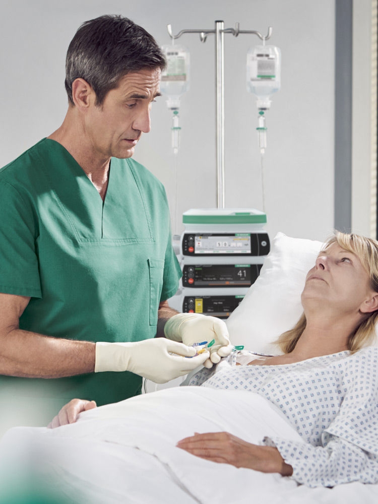 A blonde woman lies in a hospital bed. She is  connected to an infusion, a Spaceplus pump can be  seen in the background. A dark-haired doctor in a  green surgical gown stands next to the bed. He is  regulating the administration of the pain medication  by infusion and talking to the woman.