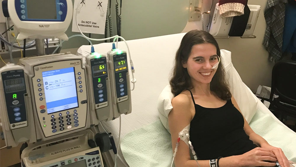 A woman with loose brown hair lies in a hospital bed and smiles into the camera. She is plugged into cables and a hospital machine to her left.