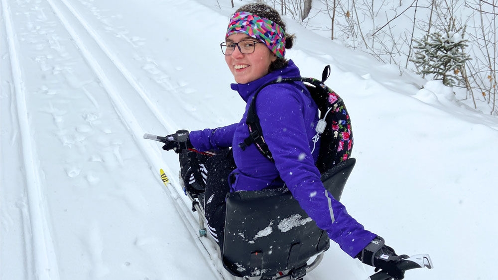 A woman looks into the camera with a smile on her face as she sits on skis and skis down a street full of snow. 