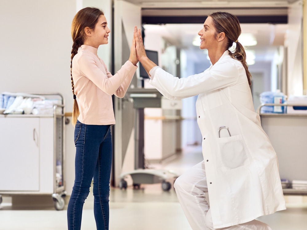 A dark-haired girl and a dark-haired doctor are  standing opposite each other in a hospital corridor,  both smiling. The doctor has crouched down to be  at eye level with the little patient. They high-five each other with both hands.