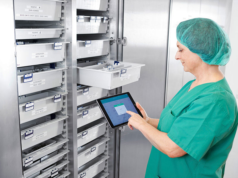 A nurse in surgical clothing holds a tablet in her hand and stands in front of a surgical cabinet