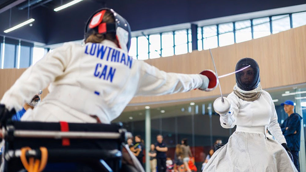 Two women practicing the sport of fencing. A sports hall and people can be seen in the background.