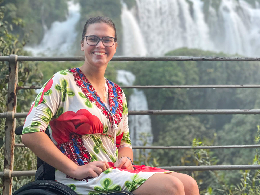 A woman with brown hair in a braid and glasses smiles at the camera, sitting in a wheelchair. Behind her is a waterfall.